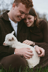 Fototapeta premium A couple in casual clothes gently petting a white goat kid outdoors, sharing a tender moment of joy in a rural setting, a scene emphasizing connection, nature, and farm life.
