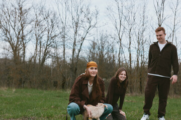 Fototapeta premium Three young adults share a playful moment with a goat kid in a grassy park, casual clothing, natural light, candid lifestyle shot capturing friendship, rural charm, and simple outdoor joy.