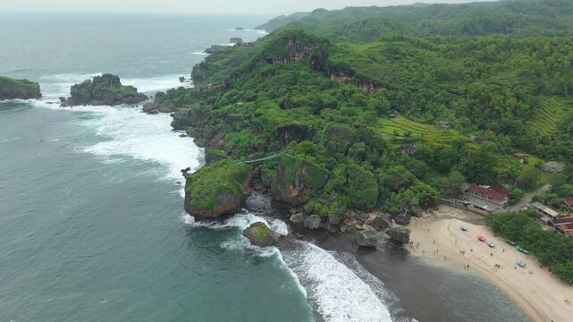 Aerial view of Siung Beach featuring rocky cliffs, a suspension bridge, lush green hills, and white waves crashing on the shore in Tepus, Special Region of Yogyakarta, Indonesia.