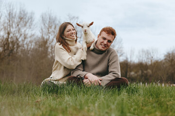 Fototapeta premium Smiling couple sitting on grass in an open field with a playful goat kid between them, capturing a warm lifestyle moment in nature and rustic charm