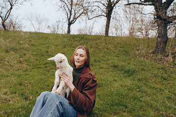 Fototapeta premium Woman sits on a grassy hill holding a white goat kid, enjoying outdoor lifestyle, gentle serene moment of companionship in a parklike countryside setting, calm and warm