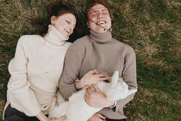 Fototapeta premium Two smiling adults lie on green grass wearing chunky sweaters, enjoying a cozy outdoor moment together with a small animal between them, showcasing lifestyle comfort, warmth, and carefree affection.