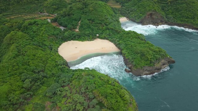 Aerial view of Sedahan Beach, a secluded sandy cove surrounded by lush green cliffs and crashing ocean waves in Girisubo, Special Region of Yogyakarta, Indonesia.
