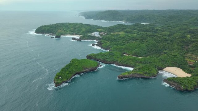 Aerial view of Sedahan Beach with its white sand and turquoise waters surrounded by lush green cliffs and crashing waves in Girisubo, Special Region of Yogyakarta, Indonesia.