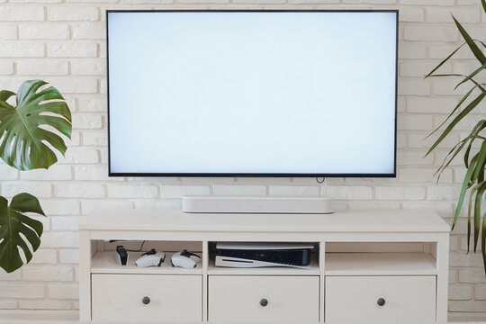 A contemporary living room scene featuring a blank television screen mounted on a white brick wall. The stylish furniture includes a light-colored TV stand with storage and potted plants, creating a s