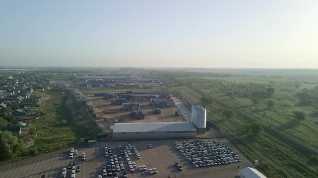 Aerial drone view of large logistics hub with container terminal, car parking lot and grain silos. Industrial supply chain and intermodal freight transport.
