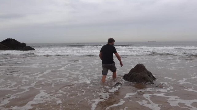 Man taking a selfie sitting on a rock in the ocean on a cloudy day