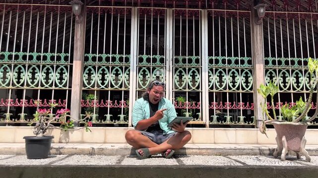 Adult asian man is sitting in front of heritage house in Kotagede while watching online worldcup championship by using an ipad
