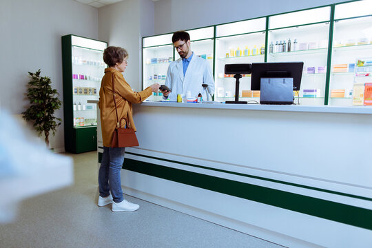 Senior woman paying for medication at modern pharmacy counter while pharmacist assists