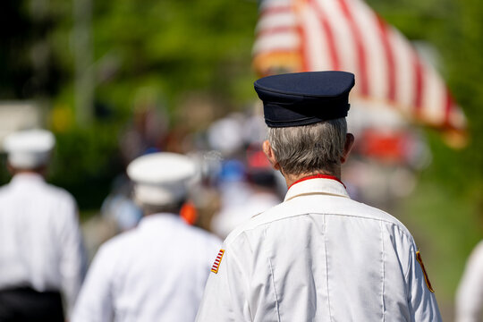 An older man in a white uniform and black cap is seen from behind at a Memorial Day or veteran's parade, American flags and what appears to be a fire truck visible ahead of him in the procession.