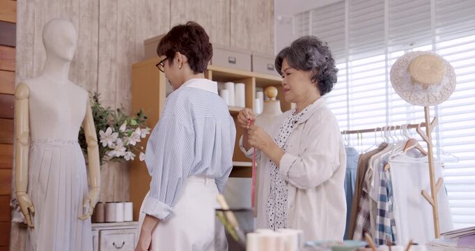 Elderly asian female fashion designer adjusting tape on shoulder of adult asian female client during fitting session boutique atelier, tailoring craftsmanship, business consultation, custom clothing