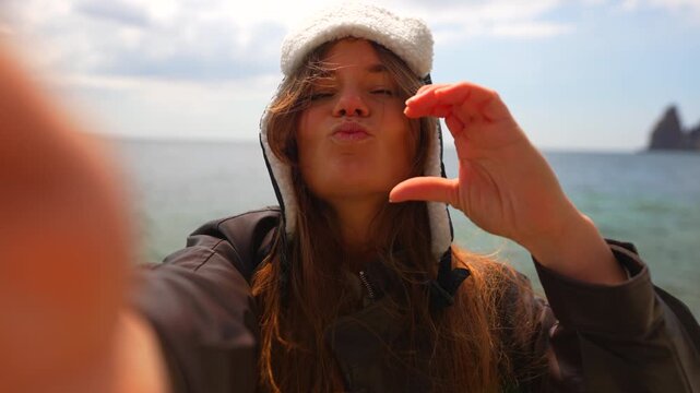 Woman beach selfie Happy young woman wearing winter hat taking a selfie while blowing kisses and making heart gestures on a sunny coastline