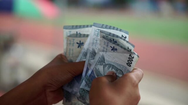 Hands counting Indonesian two thousand rupiah banknotes with a blurred outdoor background.