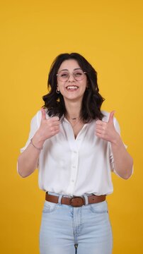 Smiling happy caucasian brunette young woman pointing up to copy space with both index fingers, then looking at camera and showing thumbs up on yellow background, vertical studio video.