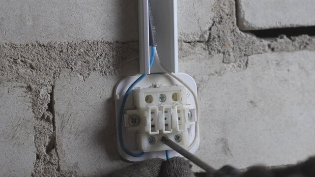 Close-up view of a professional electrician wearing protective gloves while using a screwdriver to connect wires in a terminal block on a rough brick wall during an electrical installation