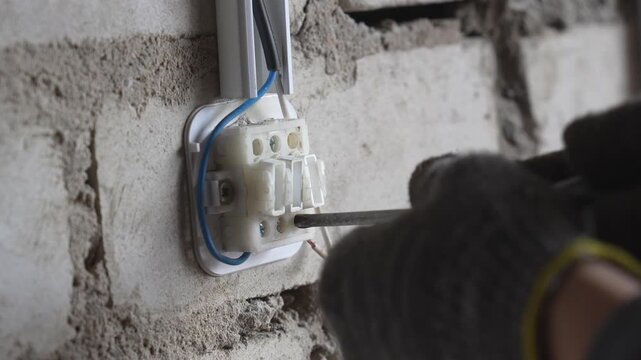 Close-up view of a professional electrician wearing protective gloves while using a screwdriver to connect wires in a terminal block on a rough brick wall during an electrical installation