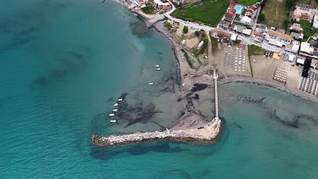 Aerial view of Roda beach pier with small boats in turquoise Ionian sea, North Corfu, Greece. Drone flight over coastal resort town with sandy beach and breakwater.