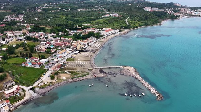 Aerial panoramic view of Roda village beach and pier in North Corfu, Greece. Mediterranean coastal resort town with clear Ionian sea, sandy shoreline, and green mountains in summer.