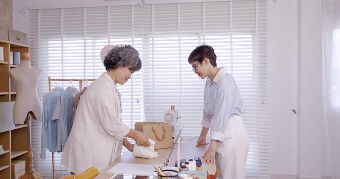 Asian senior female seamstress standing at counter handing custom garment inside paper bag to asian adult female customer inside boutique atelier small business fashion shop with sewing tools