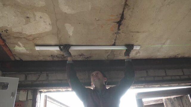 Professional electrician wearing gloves installing a new fluorescent light fixture on a rough concrete ceiling, working diligently during a building renovation project in a garage or workshop