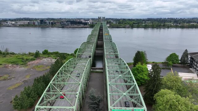 Portland Oregon Interstate Bridge with green steel arch spans crossing Columbia River, busy multi-lane highway traffic connecting Oregon and Washington state under cloudy overcast sky. Aerial flyover.