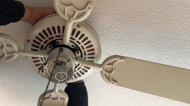 A person adjusts a light fixture on a ceiling fan, focusing on maintenance and home improvement. Suitable for DIY tutorials or home renovation content.