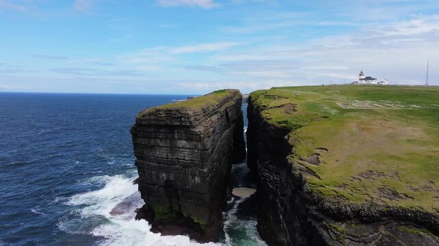 A dramatic sea stack divides the landscape at Loop Head with a distant lighthouse. Captures the rugged beauty of the Irish coast. Suitable for travel and nature content.