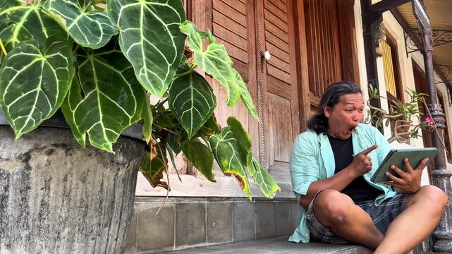 Indonesian sitting down on a Javanese house&rsquo;s terrace, watching football championship online using an ipad during the day