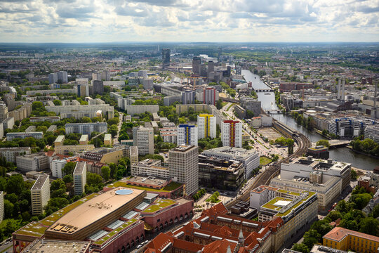 Aerial panoramic view of Berlin, Germany, with urban skyline, river, bridges, residential buildings, green parks and dramatic cloudy sky, showing the cityscape from above.