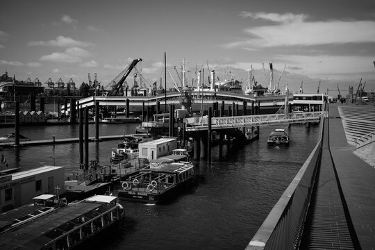Black and white panoramic view of Hamburg harbor, Germany, with waterfront promenade, boats, museum ships, modern architecture and dramatic sky in a maritime urban scene.