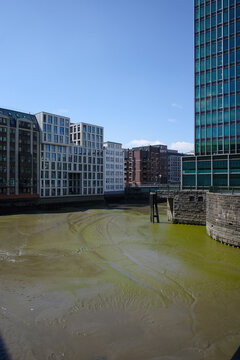 Urban waterfront view in Hamburg, Germany, showing modern and historic buildings along a tidal canal at low tide, with exposed mudflat, city architecture and clear blue sky.