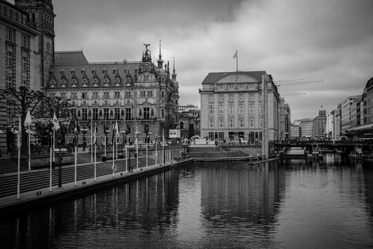 Black and white panoramic view of Hamburg City Hall and waterfront buildings along a canal, with dramatic cloudy sky, reflections and historic urban architecture in Germany.