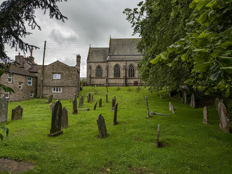 View of St Margaret's Church and its historic graveyard in Yorkshire with weathered stone headstones, lush green grass, and stone buildings in Hawes, England, United Kingdom.