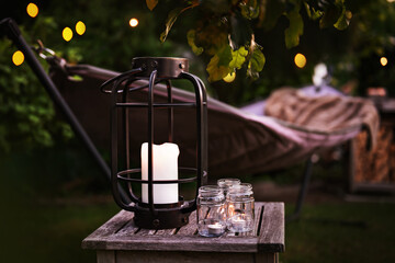 Fototapeta premium Hammock with lantern and fairy lights in evening garden setting. Cozy evening atmosphere with candlelight. Background for leisure and relaxation for outdoors.
