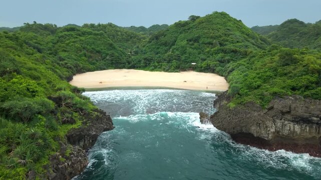 Aerial view of Sedahan Beach with white sand and turquoise waves crashing on the shore surrounded by lush green hills in Girisubo, Special Region of Yogyakarta, Indonesia.