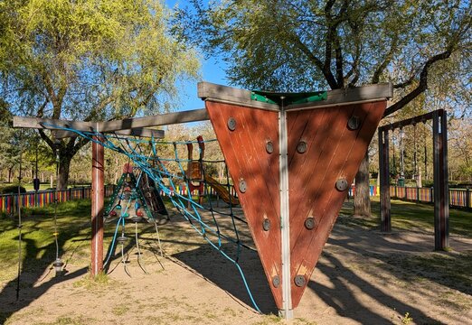 Zona de juegos infantil al aire libre, con estructura de escalada de madera, red de cuerdas, tobog&aacute;n y columpio, en jard&iacute;n de parque p&uacute;blico 