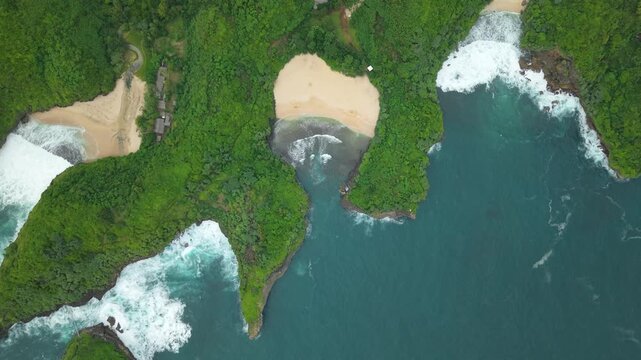 Aerial view of Sedahan Beach featuring a secluded sandy cove surrounded by lush green tropical vegetation and ocean waves in Girisubo, Special Region of Yogyakarta, Indonesia.