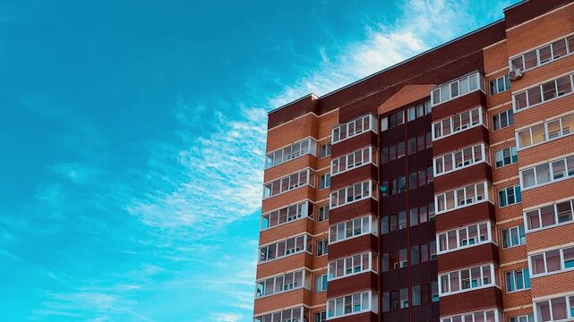 Brick apartment tower under blue sky, sunlit red facade with repeating balcony rows and window patterns, wispy clouds, crisp urban
