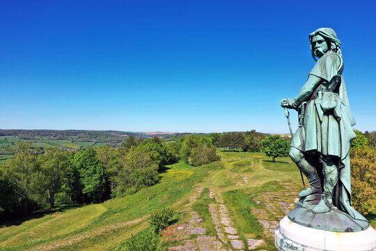 Das monumentale Vercing&eacute;torix-Denkmal auf einem steinernen Sockel, eingebettet in eine gr&uuml;ne H&uuml;gellandschaft mit B&auml;umen unter einem strahlend blauen Himmel.