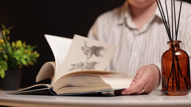 Person turning pages of an open book with reed diffuser and plant nearby
