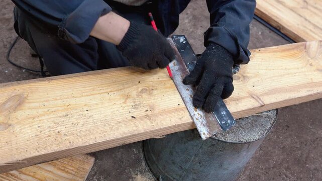 Close-up view of a professional carpenter wearing protective gloves and marking a straight line on a wooden board with a pencil and a try square before cutting it for a new project
