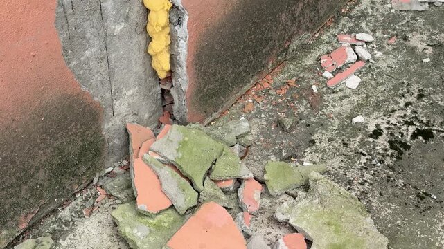 Cracked plaster corner sealed with foam, brick backdrop, vertical leak stain, damp mossy ground, scattered debris, construction