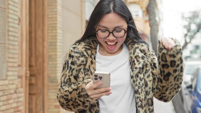 Young hispanic woman smiling at smartphone, holding device while walking on a city street; joy connection.