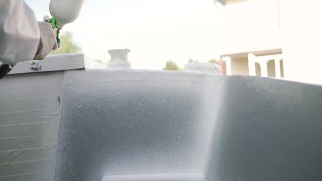 Close up of a worker in a protective suit using an airbrush spray gun. Person paints a large surface, like a boat, with white or gray paint. Outdoor renovation or repair project