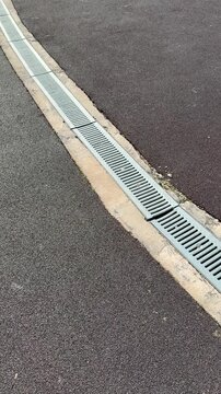 Galvanized drainage grate along stadium curb, long metal channel between asphalt and concrete, repeating slot pattern, leading