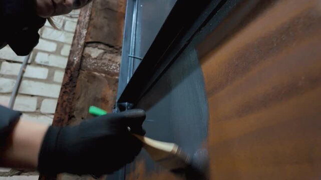 Professional worker in protective gloves carefully applying dark gray primer paint to a rusty metal surface. Preparing it for finishing and preventing corrosion during construction or renovation work