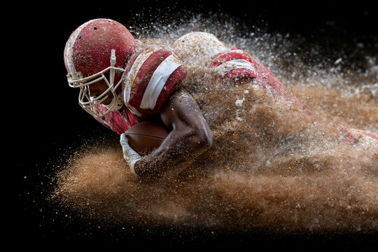Athlete breaks through dirt with fierce determination. Helmet gleams under motion, dirt flies around him. His body leans low, arms locked tight with ball