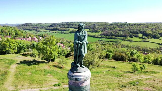 Das monumentale Vercing&eacute;torix-Denkmal auf einem steinernen Sockel, eingebettet in eine gr&uuml;ne H&uuml;gellandschaft mit B&auml;umen unter einem strahlend blauen Himmel.