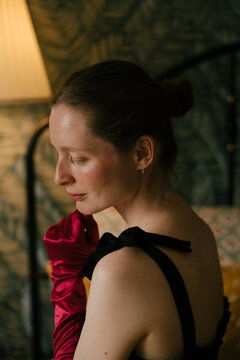 Elegant freckled woman in black dress and red gloves posing in vintage interior with soft lamp light.