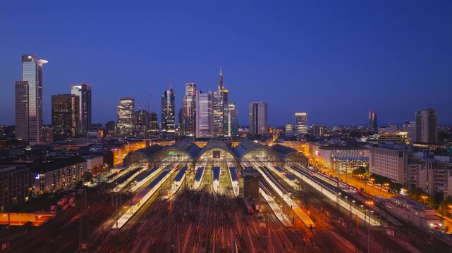 Aerial view of Hauptbahnhof train station with illuminated platforms and the modern city skyline at dusk in Frankfurt am Main, Hessen, Germany.